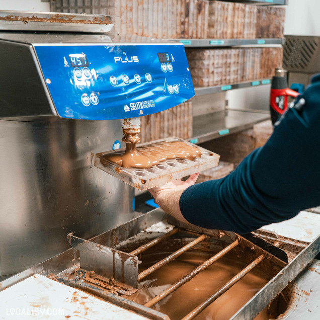 Un chocolatier remplit un moule à pralines sous une machine à tempérage de chocolat de marque Selmi, dans un atelier de fabrication artisanale.