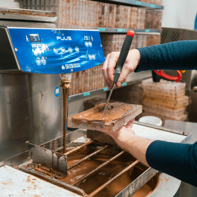 Un chocolatier égalise le chocolat fondu dans un moule à l’aide d’une spatule, devant une machine professionnelle à tempérage.