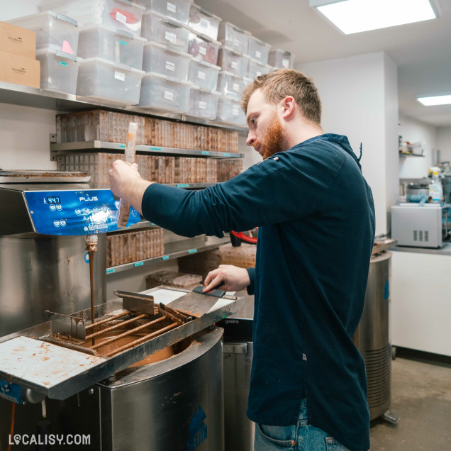 Un artisan chocolatier travaille avec concentration devant la machine à chocolat Selmi, dans un atelier bien organisé avec des étagères de moules.