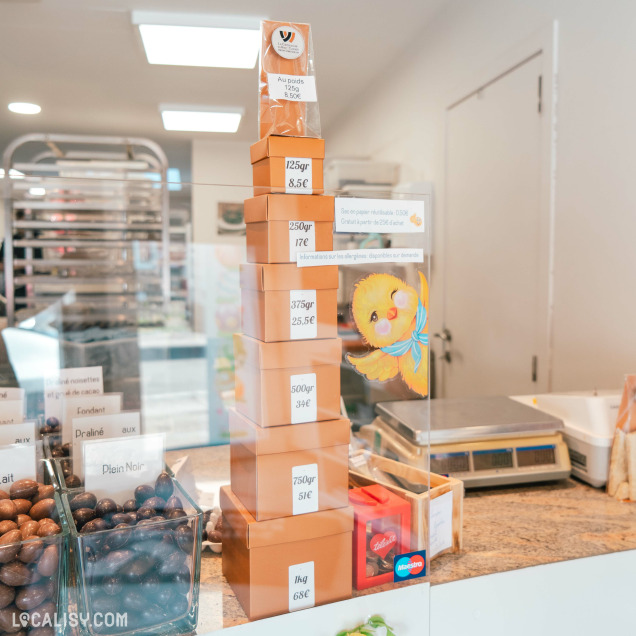 Vue de la vitrine d'une chocolaterie affichant des boîtes empilées avec les prix au poids, allant de 125g à 1kg, avec des bonbons de chocolat visibles en arrière-plan.