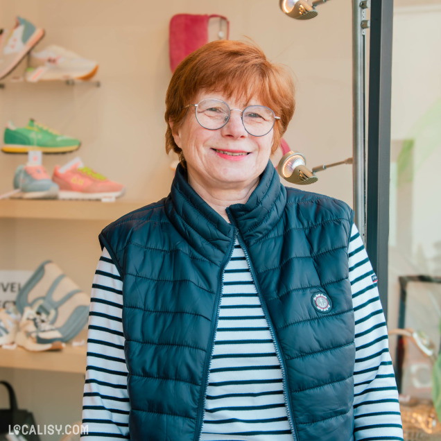 Portrait d'une femme souriante employée chez chaussures Lehane Aywaille portant une doudoune sans manches bleu marine et un haut rayé, debout à l'entrée d'un magasin de chaussures.