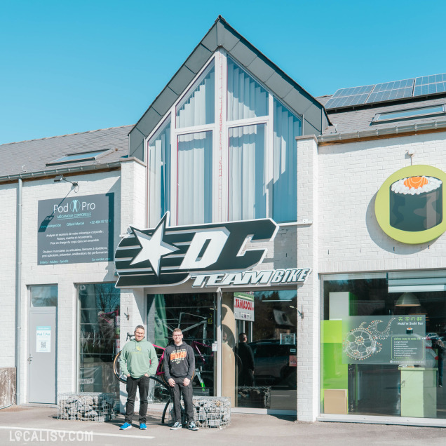 Façade extérieure du magasin DC Team Bike à côté d’un bar à sushi, avec deux personnes posant devant l’entrée sous une enseigne imposante en forme d’étoile et de silhouette de cycliste.