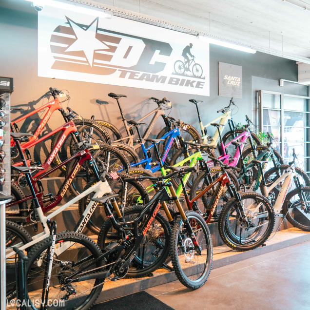 Intérieur du magasin DC Team Bike présentant un mur complet de vélos Santa Cruz suspendus, dans différentes couleurs vives comme rouge, noir, bleu et vert, avec un grand logo mural « DC Team Bike ».