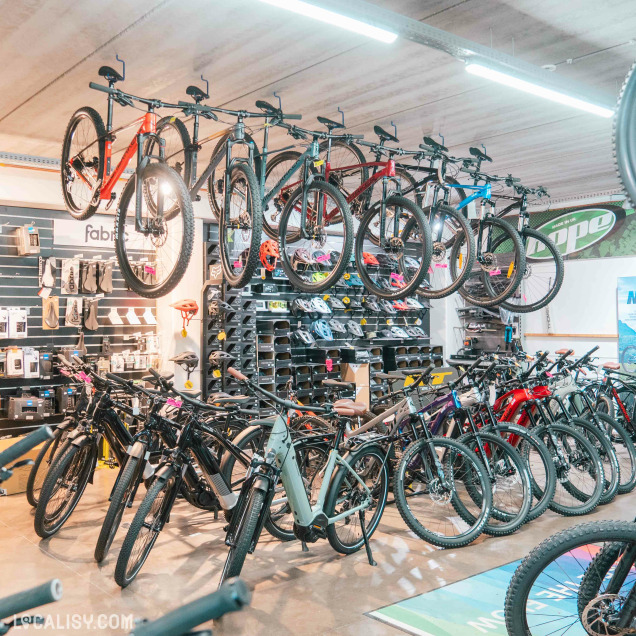 Section du magasin avec des vélos accrochés au plafond et d'autres modèles électriques en exposition au sol, avec des accessoires comme des selles, casques et outils visibles sur les étagères murales.