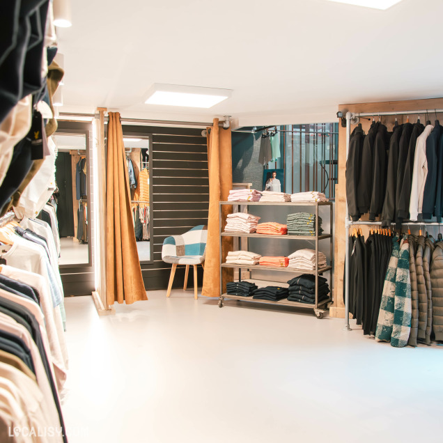 Cabines d’essayage et portants de vêtements bien alignés dans un coin cosy du magasin, avec fauteuil d’appoint et ambiance chaleureuse grâce à l’éclairage doux.