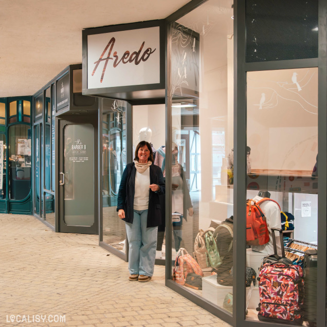 Devanture de la boutique Aredo, avec une femme souriante debout à l'entrée. La vitrine expose plusieurs sacs à dos colorés et une sélection de vêtements visibles à travers les vitrines modernes.