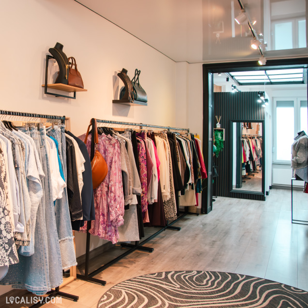 Vue d’un portant de vêtements colorés et féminins, incluant robes, vestes et manteaux, avec des sacs exposés en hauteur et une entrée lumineuse au fond du magasin.