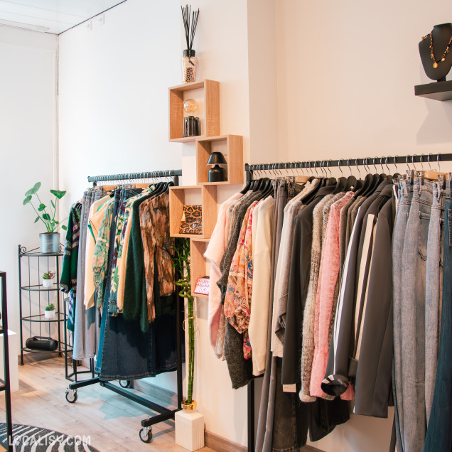 Vue latérale d’un portant avec une collection de vêtements d’automne-hiver : pulls, vestes, jeans et tissus à motifs floraux, avec une décoration murale et des plantes vertes.