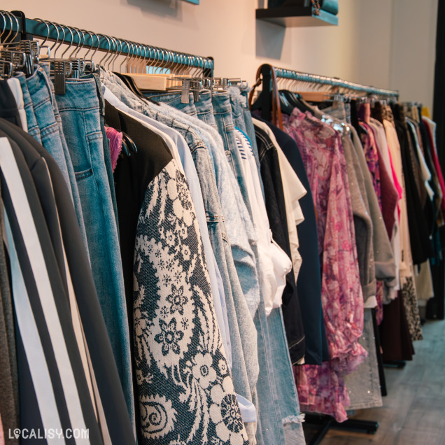 Gros plan sur une rangée de vêtements : jeans, tops brodés, robes fleuries, pulls en laine et blouses pastel, bien présentés sur cintres noirs.