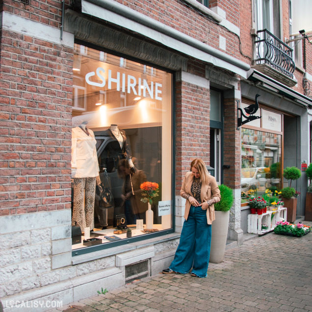 Devanture du magasin Shirine avec une grande vitrine présentant des tenues chic, et une femme debout devant, vêtue d’un blazer beige et d’un jean large, dans une rue commerçante en pavés.