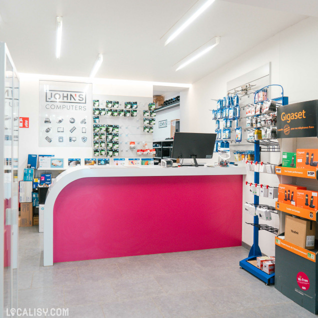 Comptoir d’accueil avec panneau “John’s Computers” à l’arrière, produits informatiques exposés au mur et sur étagères, ambiance claire et organisée.
