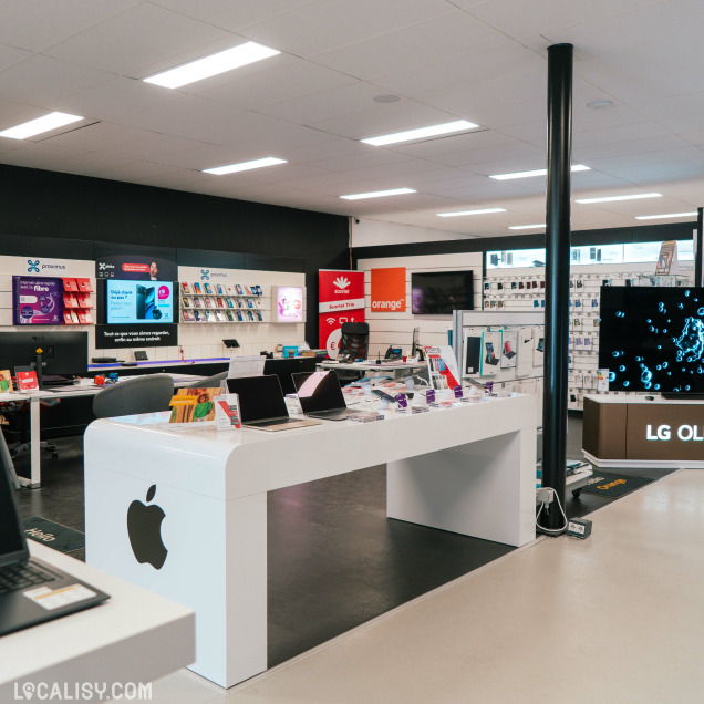 Vue d’ensemble du magasin avec un espace dédié aux produits Apple, aux télécommunications (Proximus, Scarlet, Orange), et divers ordinateurs portables en démonstration sur une table blanche brillante.