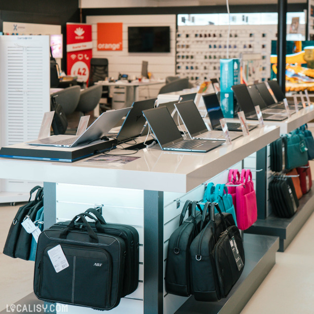 Table d’exposition avec plusieurs ordinateurs portables alignés, et un assortiment de sacs de transport colorés suspendus en dessous, dans un magasin de matériel informatique.
