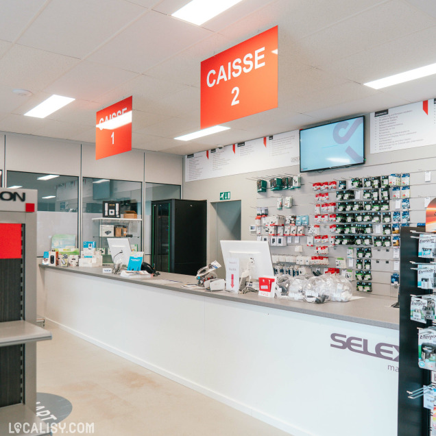 Espace caisse avec deux guichets identifiés "Caisse 1" et "Caisse 2", et un mur d’accessoires électroniques (piles, cartes mémoire, adaptateurs) derrière le comptoir.