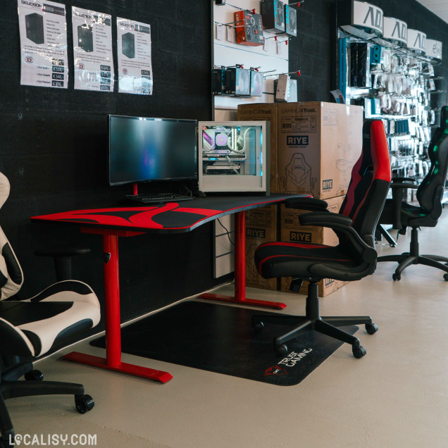 Coin gaming du magasin avec un bureau rouge et noir, un écran, un PC fixe RGB transparent, et plusieurs chaises gamer alignées le long d’un mur noir.