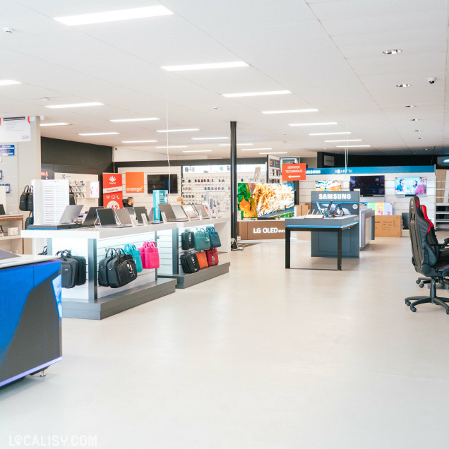 Vue panoramique du magasin avec différents espaces dédiés aux ordinateurs portables, accessoires, TV LG et Samsung. Présence de marques télécom comme Orange et Scarlet, dans un environnement lumineux et moderne.