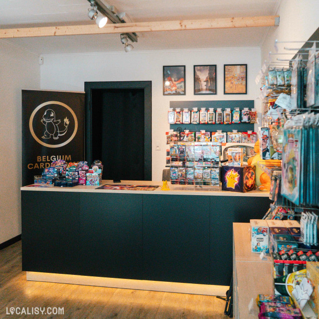 Comptoir principal d’une boutique dédiée aux cartes et produits dérivés Pokémon. Derrière le comptoir, un mur bien garni de boosters, coffrets et articles de collection sous blister. Le logo "Belgium Card Games" avec un Salamèche est visible à gauche. Des articles sont également exposés à droite sur des rayons muraux.