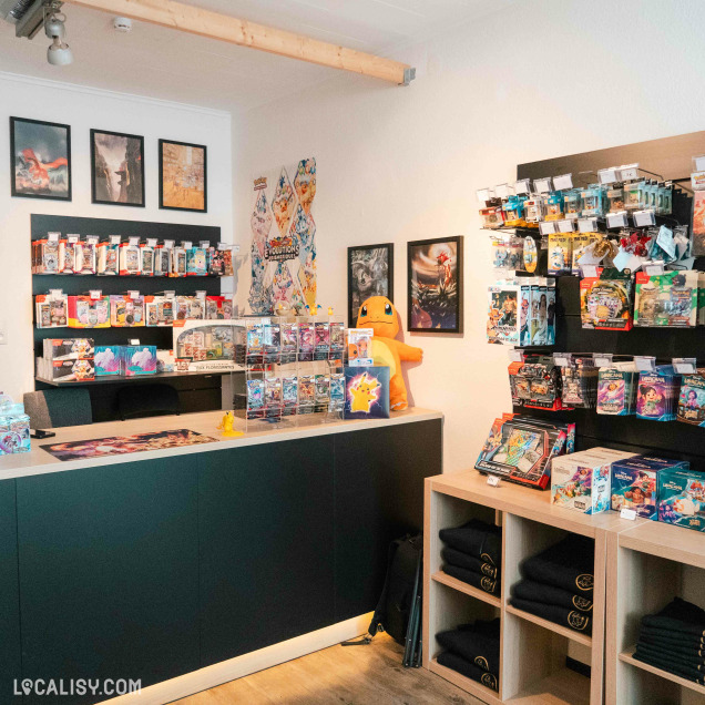 Autre angle du comptoir d’un magasin Pokémon, avec un grand peluche Salamèche en fond. Le mur du fond est orné de posters et de boosters exposés. À droite, des étagères remplies de produits Pokémon : decks, boosters, boîtes et vêtements pliés. L’ambiance est chaleureuse et organisée.