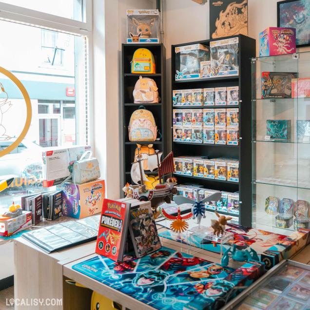 Vue intérieure d’un magasin de collection mettant en avant l’univers Pokémon. À l’avant-plan, une table avec des cartes à jouer, figurines et livres Pokémon. En arrière-plan, des étagères garnies de figurines Pop!, de sacs à dos Pokémon colorés et d’autres objets de collection. La lumière naturelle entre par une grande vitrine sur la gauche.