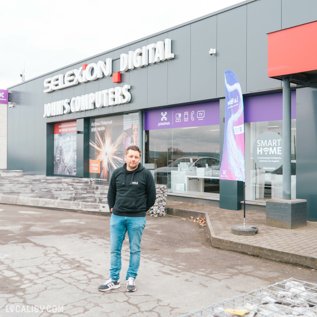 Un homme se tient debout devant la façade du magasin Selexion Digital John's Computers. Il porte un sweat noir et un jeans bleu. La devanture du commerce arbore des enseignes de Proximus, avec des visuels mettant en avant des services numériques comme l’internet ultra rapide et les solutions smart home. Le décor est moderne, avec un habillage de façade gris foncé et une banderole publicitaire colorée installée près de l’entrée.