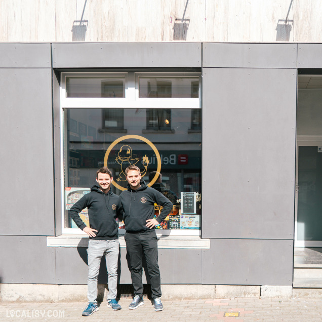 Deux hommes souriants portant des sweats à capuche noirs avec un logo, posent devant la vitrine de leur commerce. Le logo du magasin, représentant une flamme et un personnage, est visible sur la fenêtre derrière eux. L’enseigne se situe en milieu urbain.