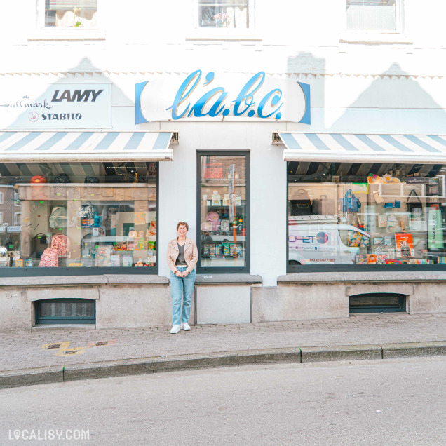 Une femme souriante pose devant la vitrine du magasin "l’a.b.c.", spécialisé en fournitures scolaires, cadeaux et papeterie. L’enseigne est décorée de stores rayés bleus et blancs, et les vitrines sont remplies d’articles colorés. La façade claire du bâtiment reflète une ambiance accueillante.
