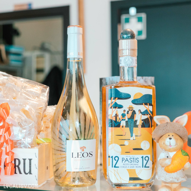 Zoom sur deux bouteilles en vente à la fromagerie : un vin blanc Leos et une bouteille de Pastis 12, entourées de paniers gourmands. L’arrière-plan laisse deviner l’univers chaleureux de la boutique.