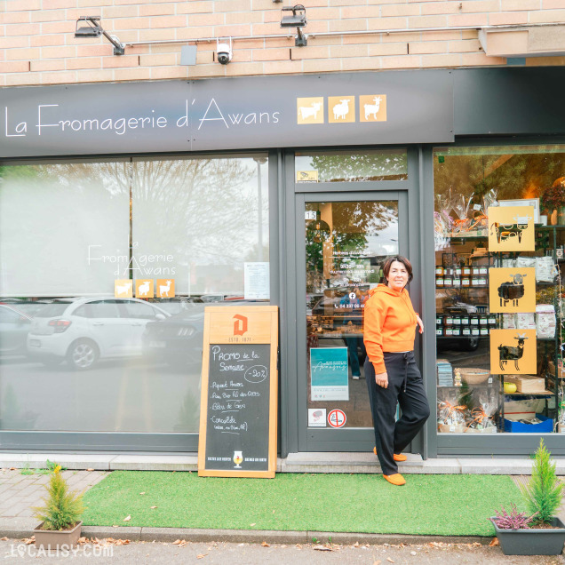 Portrait d’une femme souriante, vêtue d’un pull orange, posant devant l’entrée de La Fromagerie d’Awans. La vitrine du magasin laisse entrevoir les produits proposés et un tableau en bois annonce les promotions.