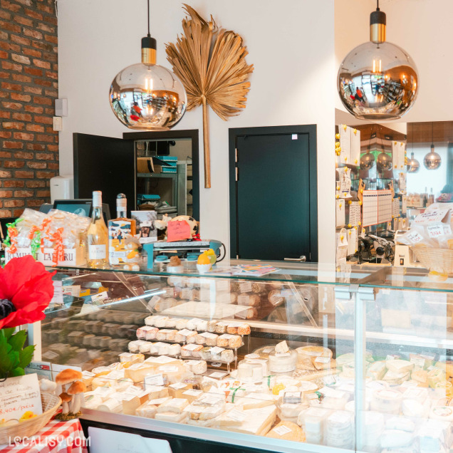 Vue intérieure de La Fromagerie d’Awans avec un grand comptoir réfrigéré rempli de fromages variés. L’ambiance est chaleureuse avec des décorations soignées, une feuille dorée accrochée au mur et des suspensions en forme de sphères métalliques.