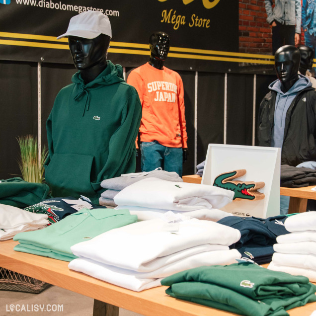 Présentation de vêtements Lacoste et Superdry sur mannequins et tables au centre du magasin.