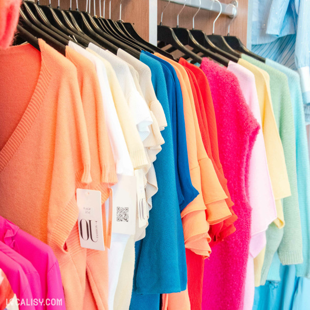Magasin de vêtements Other Side à Amay présentant des pulls colorés suspendus sur cintres noirs.