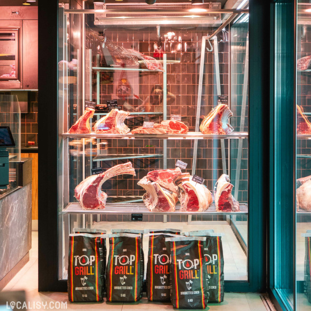 Vitrine réfrigérée de la boucherie Debruge - Halle de l'Artisan à Chênée présentant des pièces de viande et des sacs de charbon.