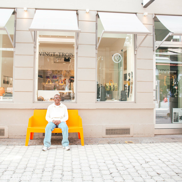 Façade du magasin de dépôt-vente Vingt-Trois Shop à Liège avec une personne assise sur un banc orange devant l'entrée.