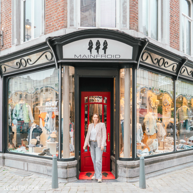 Façade extérieure de la Boutique Mainfroid à Huy, un magasin de vêtements pour hommes, avec sa vitrine et une personne à l'entrée.