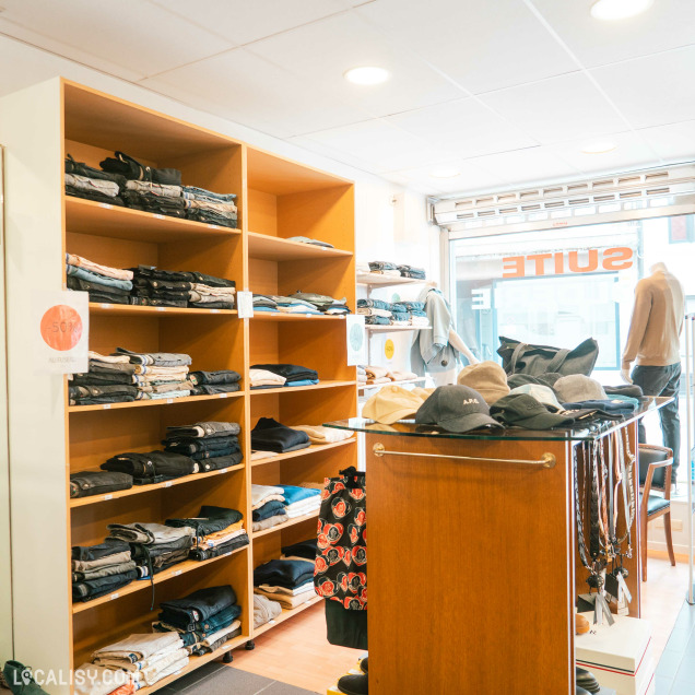Vue d'ensemble de l'intérieur du magasin de déstockage Au Fuseau Suite à Spa, avec des vêtements et des accessoires.