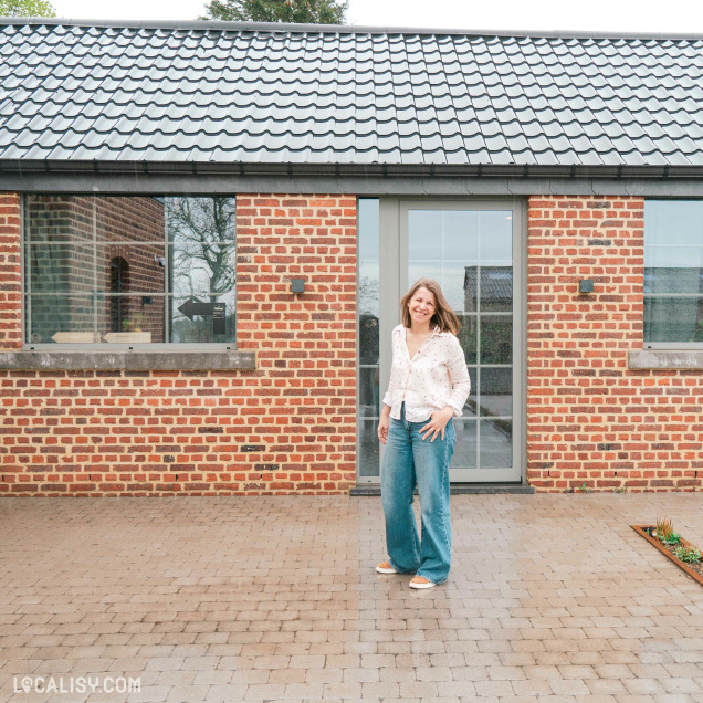 Une femme souriante se tient devant un bâtiment en briques rouges rénové avec de grandes baies vitrées, dans une ambiance chaleureuse et authentique.