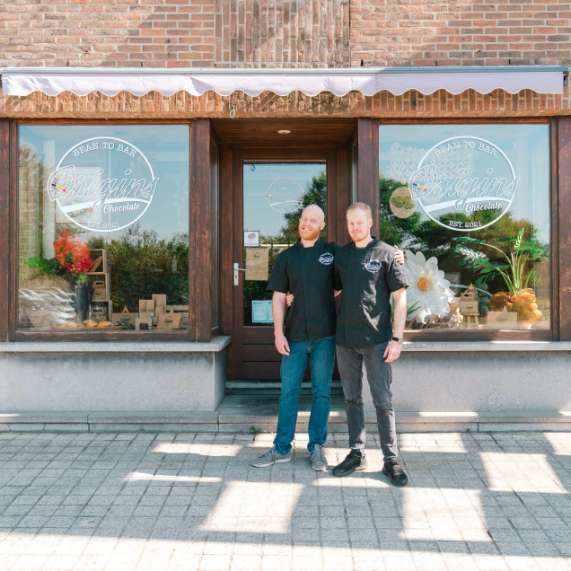 Devant leur boutique artisanale de chocolat "Origins", deux chocolatiers posent fièrement, complices et souriants, dans une ambiance chaleureuse et authentique.