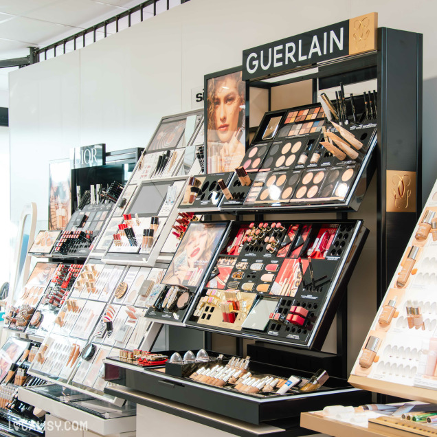 Présentoirs de maquillage Guerlain et Dior bien agencés, exposant fonds de teint, palettes de fards et rouges à lèvres dans une boutique élégante.