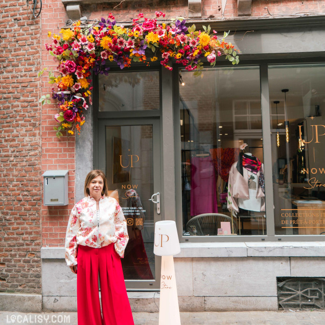 Façade du magasin de vêtements Up now à Huy, avec une femme posant devant l'entrée décorée de fleurs.