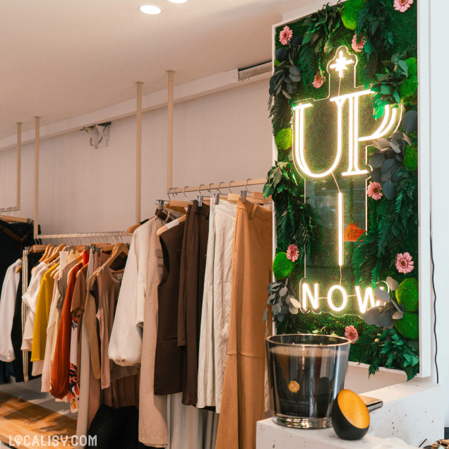Intérieur du magasin de vêtements Up now à Huy, avec un néon décoratif sur un mur végétal.