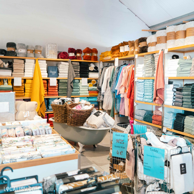 Magasin de linge de maison avec des étagères remplies de serviettes colorées, peignoirs et textiles, ainsi que des paniers et bacs promotionnels au centre.